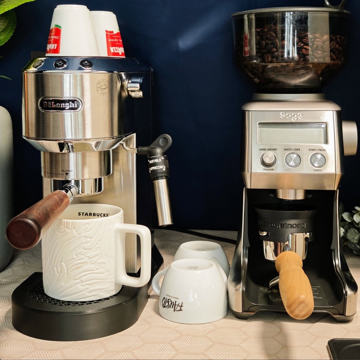 De'Longhi Dedica color argento sulla vaschetta raccogligocce Honeycomb, una tazza sotto, accanto un macinacaffè con l'imbuto dosatore inserito, davanti a una parete blu scuro